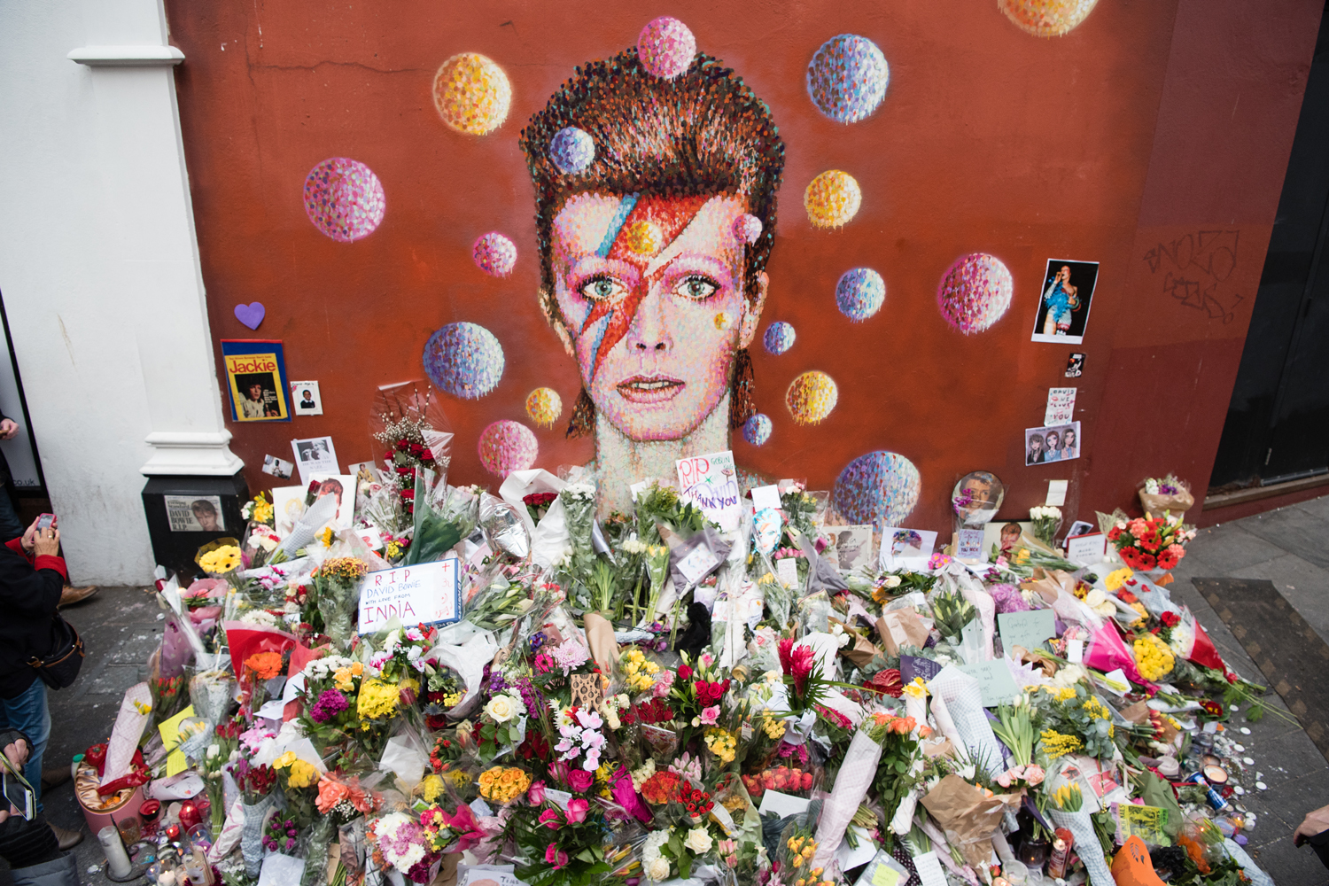 Floral tributes are seen beneath a mural of British singer David Bowie, painted by Australian street artist James Cochran, aka Jimmy C, the day after the announcement of Bowie's death, in Brixton, south London, on January 12, 2016. Music legend David Bowie was famously private during his lifetime -- and in death, as a string of questions about the circumstances of his passing remained unanswered. His official social media accounts had announced the shock news of his death at 69 on January 11, 2016: "David Bowie died peacefully today surrounded by his family after a courageous 18-month battle with cancer," adding a request for privacy for the grieving family. / AFP / LEON NEAL